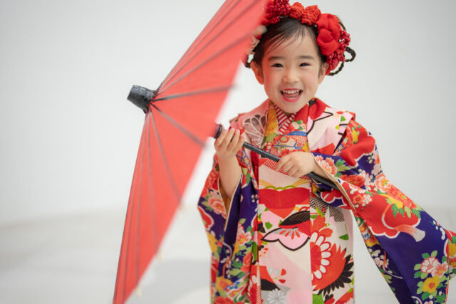 広島市安佐南区・十色写真STUDIOで撮影した7歳の七五三。色鮮やかな着物姿の女の子が赤い和傘を肩にかけ、自然な笑顔を見せている