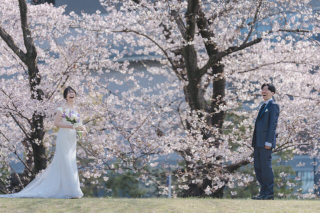 満開の桜の下でブーケを持つ花嫁と離れて立つ新郎｜広島の桜ロケーション前撮り